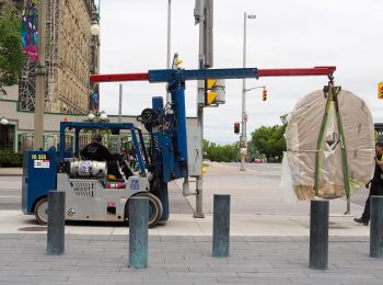 Photograph, colour; city street, small lift truck with a large, flat oval stone hanging from its boom.