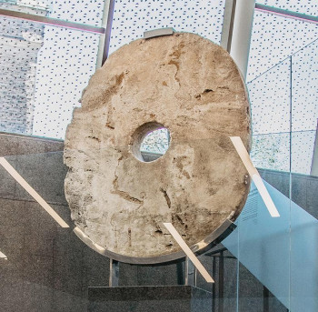 Large flat stone, carved into an oval with a hole in its centre on a stand in a museum.