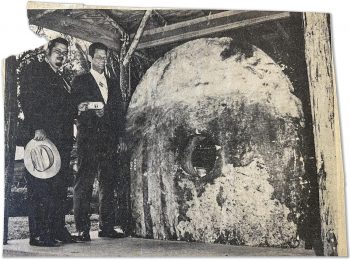 Newspaper clipping, photo, black and white; two men in suits on a platform under a small roof in front of a tall, oval stone disc with a hole carved in the middle.