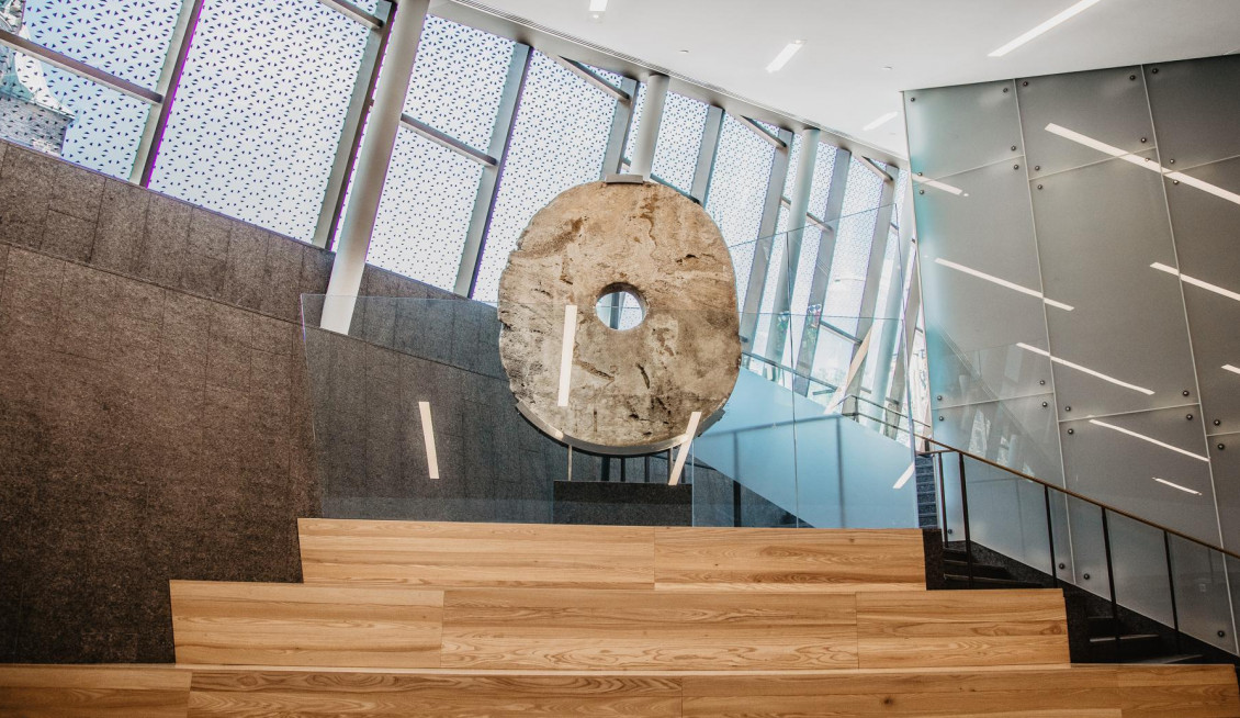 Large flat stone, carved into an oval shape with a hole in its centre on a stand at the top of a set of broad, wooden stairs in an atrium.